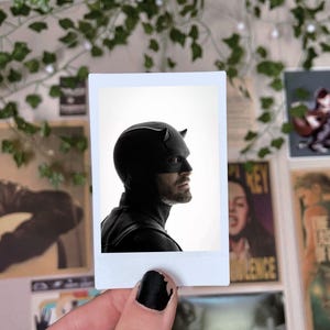 May include: A square instant film photograph featuring a profile view of a person wearing a black Daredevil mask with small horns. The subject has a visible beard and is set against a bright white background, captured in a high-contrast style.