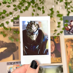 May include: A Polaroid photo of a character from a video game. The character is a humanoid alien with green skin and a blue helmet. The character is wearing a dark blue suit with a light blue stripe down the front.