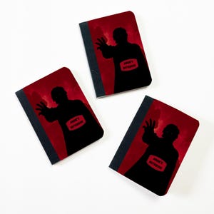 May include: Three notebooks with a red background and a black silhouette of a person reaching out. Each notebook has the text "Angie's Notebook" on a red rectangle. The spine of each notebook is dark gray.