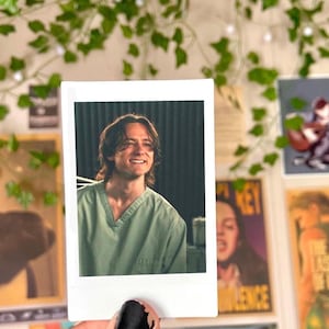 May include: A polaroid-style photograph featuring a smiling person wearing a green scrub top. The photo is held against a backdrop of decorative elements, including posters and artificial greenery.