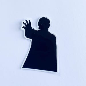 May include: A black silhouette sticker of a person in a long coat, with their arm outstretched and hand raised. The sticker has a white border and is on a white background. The person's hair is visible.