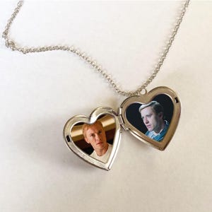 Puede incluir: Un collar con un relicario plateado en forma de corazón, abierto para revelar dos fotografías en color. El relicario cuelga de una delicada cadena. Las fotos muestran a dos hombres. El relicario está sobre una superficie blanca.