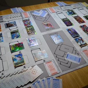 May include: A tabletop game setup featuring a playmat with designated areas for cards. Various colorful trading cards are arranged on the mat, along with stacks of cards and card sleeves. The game appears to be in progress.