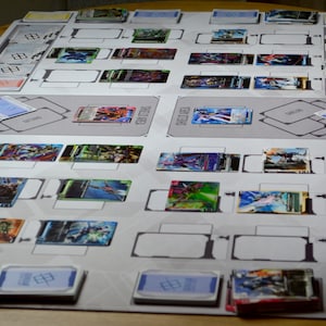 May include: A tabletop card game setup with a gray playmat featuring designated areas. Various colorful trading cards are arranged on the mat, along with stacks of cards and card holders. The cards display vibrant illustrations.