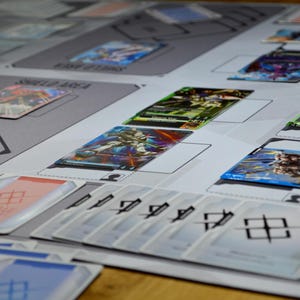 May include: A tabletop game setup featuring various trading cards and playmats. The cards display colorful illustrations of characters and symbols. The playmats have designated areas for cards, with text labels like "SHIELD AREA".