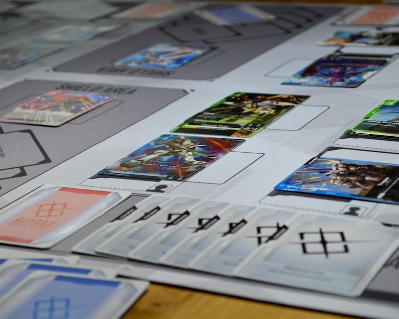 May include: A tabletop game setup featuring various trading cards and playmats. The cards display colorful illustrations of characters and symbols. The playmats have designated areas for cards, with text labels like "SHIELD AREA".