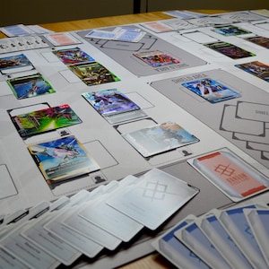 May include: A gray playmat with a grid design, featuring various trading cards with colorful illustrations. Several stacks of cards are visible, along with cards placed in designated areas labeled "Shield Area" and "Shields". The cards have a futuristic theme.