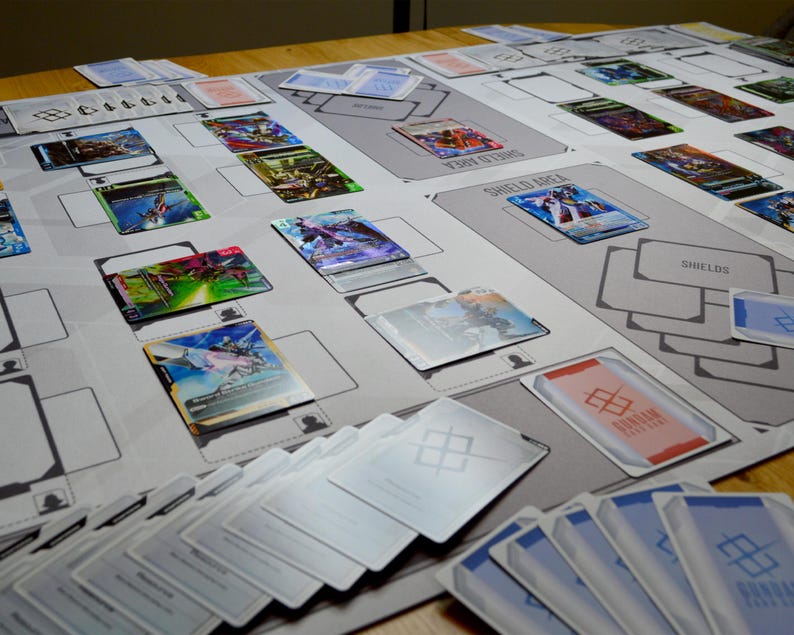 May include: A gray playmat with a grid design, featuring various trading cards with colorful illustrations. Several stacks of cards are visible, along with cards placed in designated areas labeled "Shield Area" and "Shields". The cards have a futuristic theme.