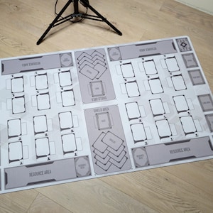 May include: A rectangular, light gray game mat with a white geometric design. It features designated areas for cards, including "Resource Area" and "Shield Area" text. The mat is designed for tabletop card games.