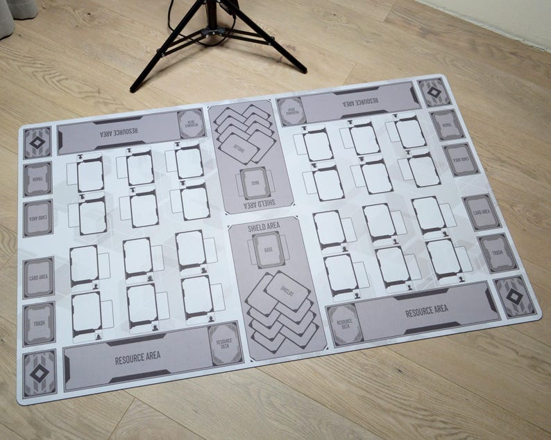 May include: A rectangular, light gray game mat with a white geometric design. It features designated areas for cards, including "Resource Area" and "Shield Area" text. The mat is designed for tabletop card games.