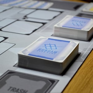 May include: Stacks of Gundam card game cards with blue and white designs, resting on a gray game mat. The mat features various game spaces and text, with a wooden table in the background. The card game's logo is visible on the card faces.