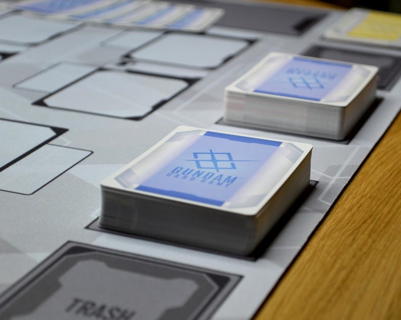 May include: Stacks of Gundam card game cards with blue and white designs, resting on a gray game mat. The mat features various game spaces and text, with a wooden table in the background. The card game's logo is visible on the card faces.