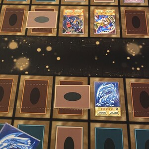 Puede incluir: Primer plano de un tablero de juego con cartas dispuestas. Las cartas son del juego de cartas coleccionables Yu-Gi-Oh! Las cartas est&aacute;n boca abajo excepto dos cartas que est&aacute;n boca arriba. Las cartas boca arriba son "Blue-Eyes White Dragon" y "Dark Magician Girl".