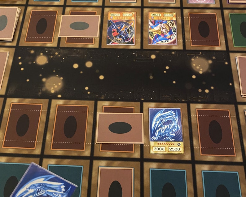 Puede incluir: Primer plano de un tablero de juego con cartas dispuestas. Las cartas son del juego de cartas coleccionables Yu-Gi-Oh! Las cartas est&aacute;n boca abajo excepto dos cartas que est&aacute;n boca arriba. Las cartas boca arriba son "Blue-Eyes White Dragon" y "Dark Magician Girl".