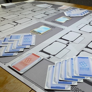 May include: A Gundam card game playmat with various zones and card layouts. Several Gundam cards are scattered across the mat, featuring blue and red accents. The playmat is gray with white and black details.