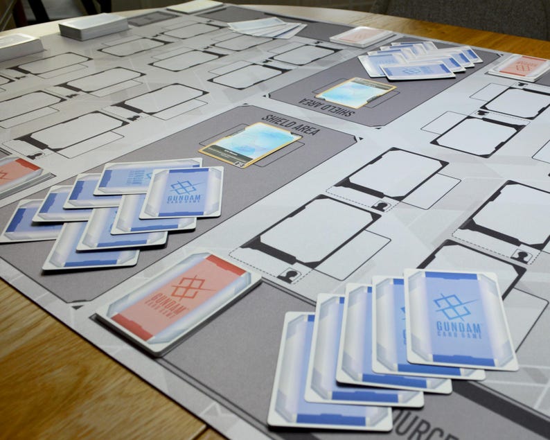 May include: A Gundam card game playmat with various zones and card layouts. Several Gundam cards are scattered across the mat, featuring blue and red accents. The playmat is gray with white and black details.