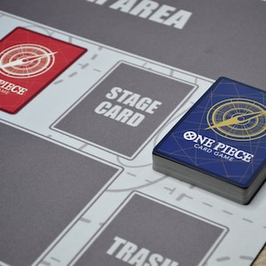 K&ouml;nnte beinhalten: Eine grau-wei&szlig;e Spielmatte f&uuml;r das One Piece Card Game mit ausgewiesenen Bereichen zum Spielen. Ein Stapel Karten mit blauem R&uuml;cken und dem Logo des Spiels liegt auf der Matte. Eine rote Karte mit dem Spiel-Logo ist ebenfalls sichtbar.
