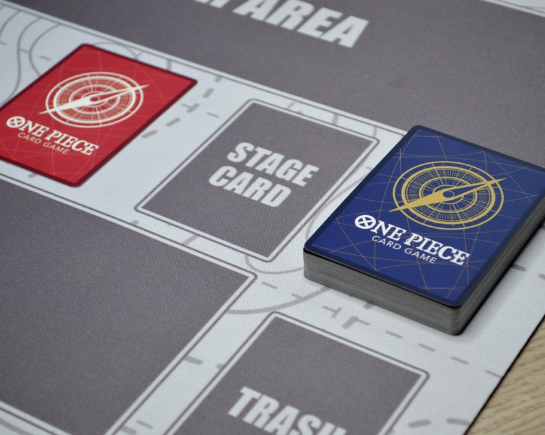 K&ouml;nnte beinhalten: Eine grau-wei&szlig;e Spielmatte f&uuml;r das One Piece Card Game mit ausgewiesenen Bereichen zum Spielen. Ein Stapel Karten mit blauem R&uuml;cken und dem Logo des Spiels liegt auf der Matte. Eine rote Karte mit dem Spiel-Logo ist ebenfalls sichtbar.