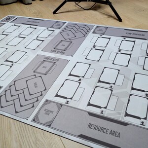 May include: A large, rectangular game mat with a gray and white design. The mat features labeled areas for resources, shields, and decks, with numerous card spaces. The overall design is geometric and organized, suitable for tabletop gaming.