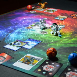 May include: A colorful game mat with a hexagonal pattern and various game pieces. Several cards are scattered around the mat, along with small, detailed figurines. The mat's background transitions through a spectrum of colors.