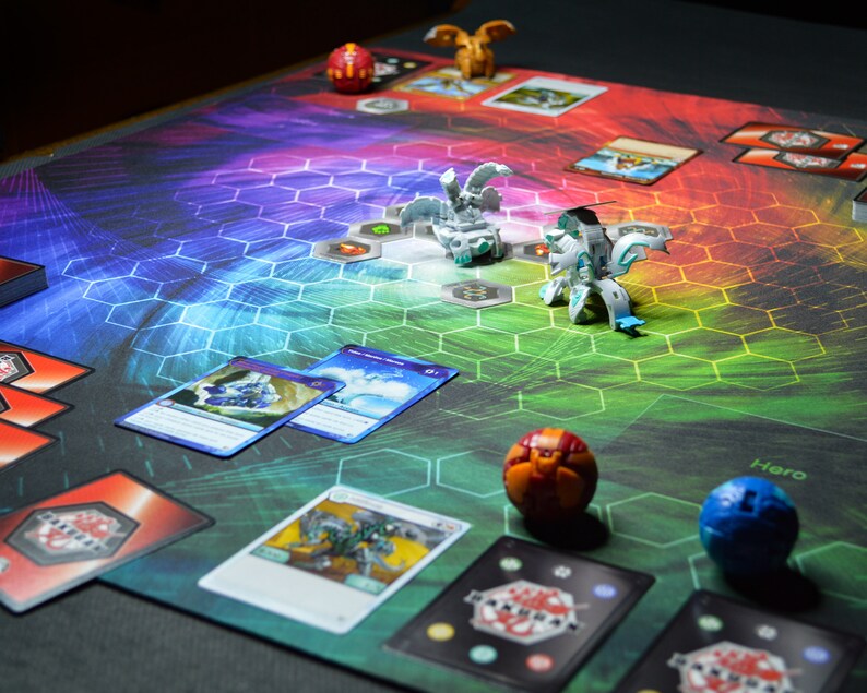 May include: A colorful game mat with a hexagonal pattern and various game pieces. Several cards are scattered around the mat, along with small, detailed figurines. The mat's background transitions through a spectrum of colors.
