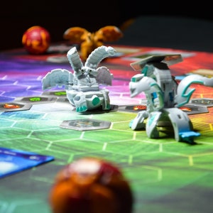 May include: A colorful game board with hexagonal spaces and two transforming toy figures. The figures are white and blue, with intricate designs. A red and orange ball is in the foreground.