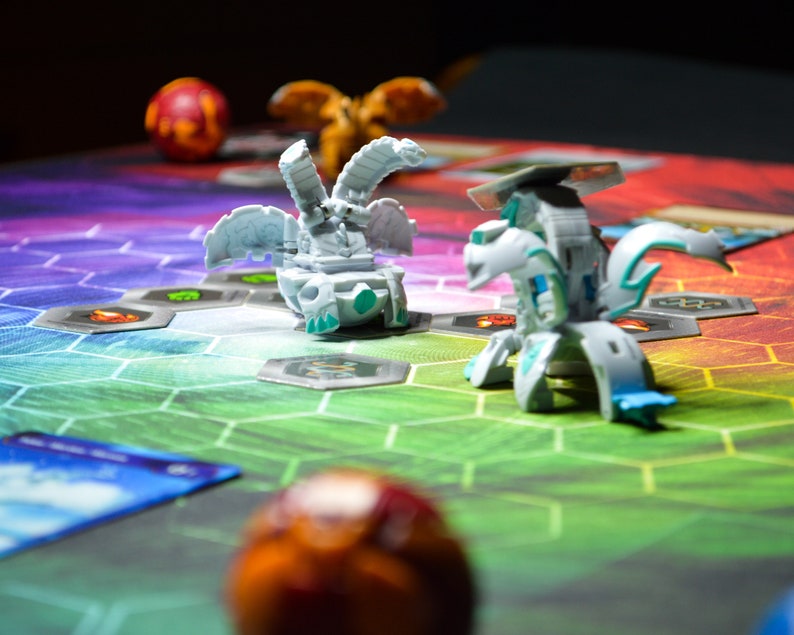 May include: A colorful game board with hexagonal spaces and two transforming toy figures. The figures are white and blue, with intricate designs. A red and orange ball is in the foreground.