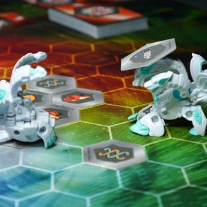 May include: Two white and teal Bakugan figures are positioned on a colorful hexagonal game board. One figure is in a battle stance, while the other is in a rolling position. A stack of cards is visible in the background.