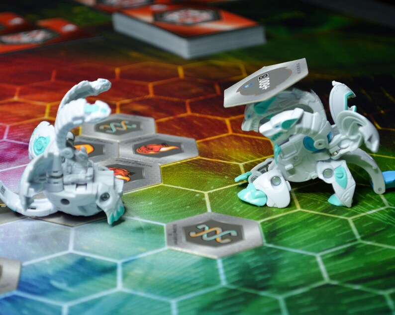 May include: Two white and teal Bakugan figures are positioned on a colorful hexagonal game board. One figure is in a battle stance, while the other is in a rolling position. A stack of cards is visible in the background.