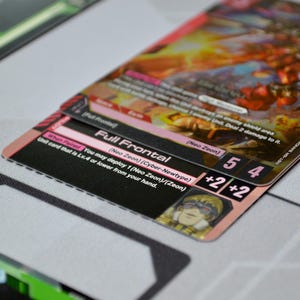 May include: Close-up of a trading card with the text "Full Frontal" and "Neo Zeon". The card has a pink and black border with the numbers 5 and 4, and a +2/+2 symbol. The background is a light gray surface.
