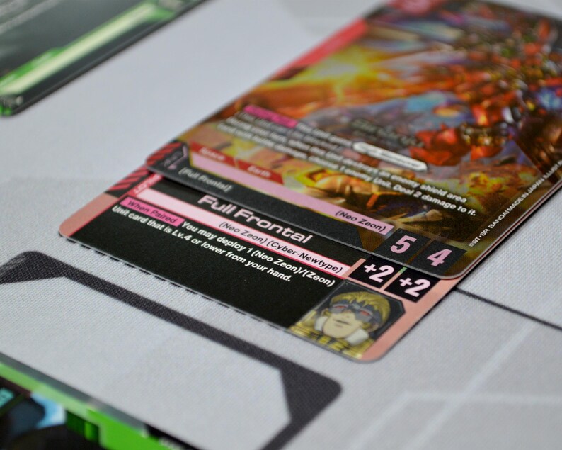 May include: Close-up of a trading card with the text "Full Frontal" and "Neo Zeon". The card has a pink and black border with the numbers 5 and 4, and a +2/+2 symbol. The background is a light gray surface.