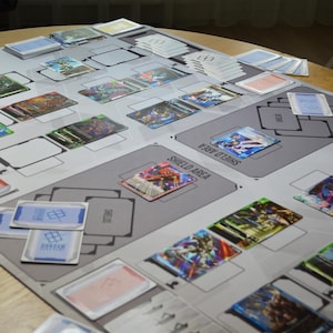 May include: A tabletop game setup featuring a gray playmat with designated areas and the text "SHIELD AREA." Various colorful trading cards are arranged on the mat, along with stacks of cards. The cards display vibrant illustrations.