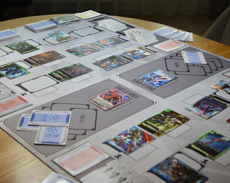 May include: A tabletop game setup featuring a gray playmat with designated areas and the text "SHIELD AREA." Various colorful trading cards are arranged on the mat, along with stacks of cards. The cards display vibrant illustrations.
