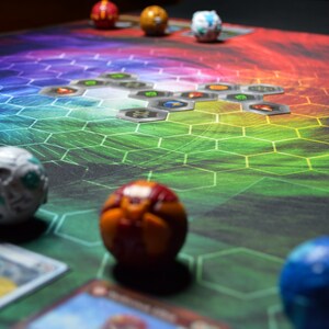May include: A colorful game board with a hexagonal pattern and several spherical game pieces. The board transitions from green to purple, with various game cards and metallic-looking tiles scattered across it. Several spherical toys are visible.