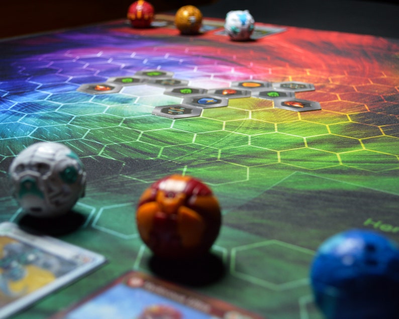 May include: A colorful game board with a hexagonal pattern and several spherical game pieces. The board transitions from green to purple, with various game cards and metallic-looking tiles scattered across it. Several spherical toys are visible.