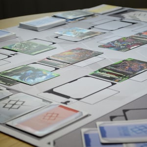 May include: A tabletop game setup featuring a playmat with designated zones and various trading cards. The cards display colorful artwork and text, with stacks of cards visible. The playmat is light gray with black borders.