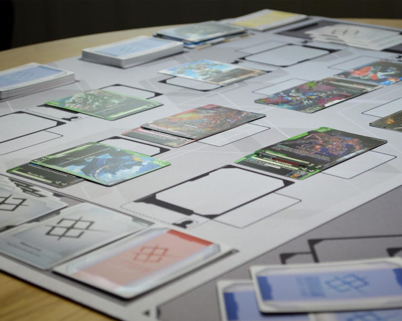 May include: A tabletop game setup featuring a playmat with designated zones and various trading cards. The cards display colorful artwork and text, with stacks of cards visible. The playmat is light gray with black borders.