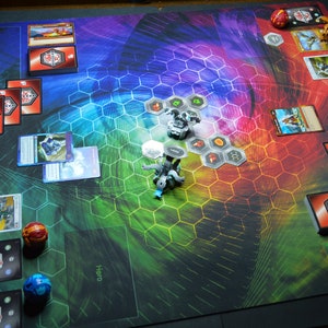 May include: A colorful game mat with a hexagonal pattern, featuring cards, game pieces, and dice. The mat has areas labeled "Energy Zone" and "Hero". The game pieces are small, detailed figures. The color scheme includes red, blue, green, and yellow.
