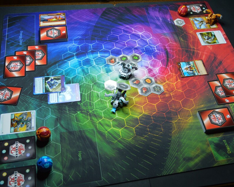 May include: A colorful game mat with a hexagonal pattern, featuring cards, game pieces, and dice. The mat has areas labeled "Energy Zone" and "Hero". The game pieces are small, detailed figures. The color scheme includes red, blue, green, and yellow.