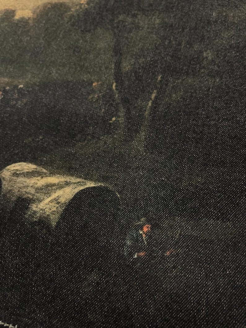 May include: A close-up of a dark denim fabric with a faded image of a landscape scene. The image appears to be of a forest with a large tree in the background and a small figure in the foreground.