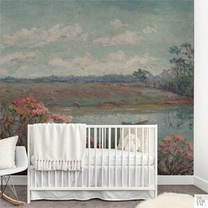 May include: A white crib with a soft white blanket and a decorative pillow. The crib is set against a large wall mural of a landscape painting with a lake, trees, and a cloudy sky. A white rocking chair is to the left.