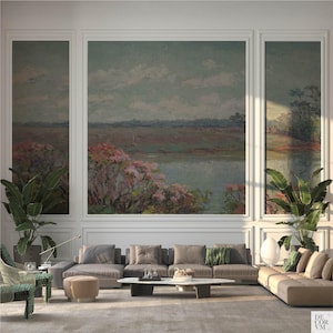 May include: A living room with a large landscape painting above a sectional sofa. The painting depicts a body of water with pink and green foliage. The room features large potted plants, a coffee table, and a patterned armchair.