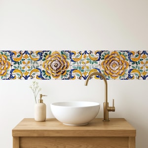 May include: A bathroom vanity with a white ceramic sink, gold faucet, and a beige soap dispenser. Above the sink is a decorative border of floral tiles in yellow, blue, and green. The vanity is made of light-colored wood.