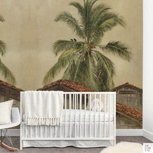 Puede incluir: Una cuna blanca con una manta blanca y un peluche frente a un mural de papel pintado. El mural representa una escena tropical con palmeras, tejados de tejas rojas y una paleta de colores apagados. Una mecedora blanca est&aacute; a la izquierda.