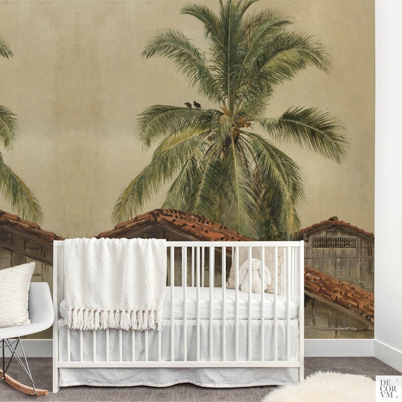 Puede incluir: Una cuna blanca con una manta blanca y un peluche frente a un mural de papel pintado. El mural representa una escena tropical con palmeras, tejados de tejas rojas y una paleta de colores apagados. Una mecedora blanca est&aacute; a la izquierda.