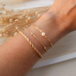Puede incluir: Un conjunto de cuatro pulseras de oro en una muñeca. Las pulseras están superpuestas, con una cadena de cuerda, una cadena delicada con pequeñas cuentas, una cadena delicada con una sola perla y una cadena con eslabones más grandes.