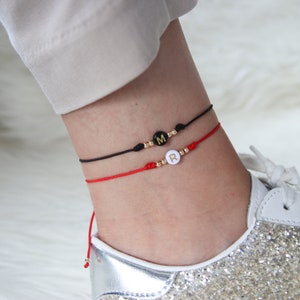 Könnte beinhalten: Zwei Fußkettchen mit schwarzem und rotem Band. Das schwarze Armband hat eine schwarze Perle mit einem goldenen Buchstaben "M" und das rote Armband hat eine weiße Perle mit einem goldenen Buchstaben "R".