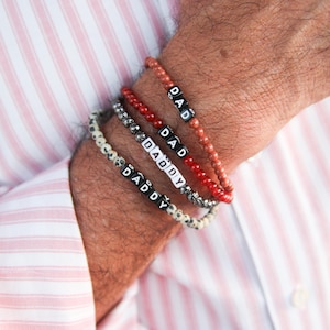 May include: Four beaded bracelets spelling "DAD" and "DADDY" in black and white letter beads. The bracelets are red, speckled, and white. The bracelets are worn on a person's wrist, with a pink and white striped shirt in the background.