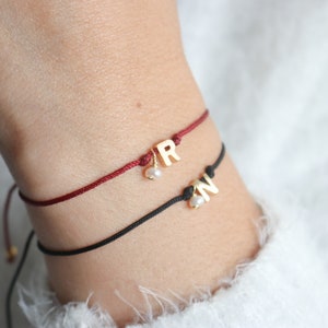 May include: Two friendship bracelets with gold letter charms. The top bracelet is red with a gold "R" charm and a small pearl. The bottom bracelet is black with a gold "N" charm and a small pearl.