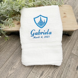 May include: White towel with blue embroidered city skyline and shield design. The text "Gabriela March 4, 2023" is embroidered below the design.